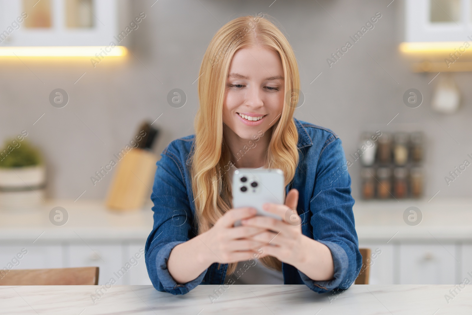 Photo of Smiling woman using smartphone at white table in kitchen