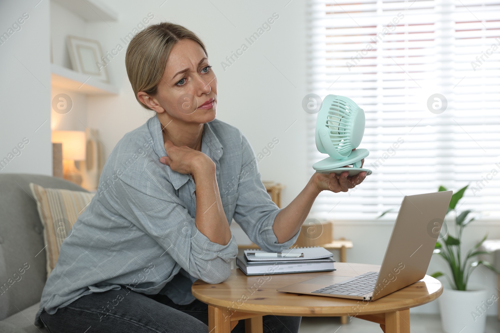 Photo of Menopause. Woman using electric fan to cool herself during hot flash at table indoors