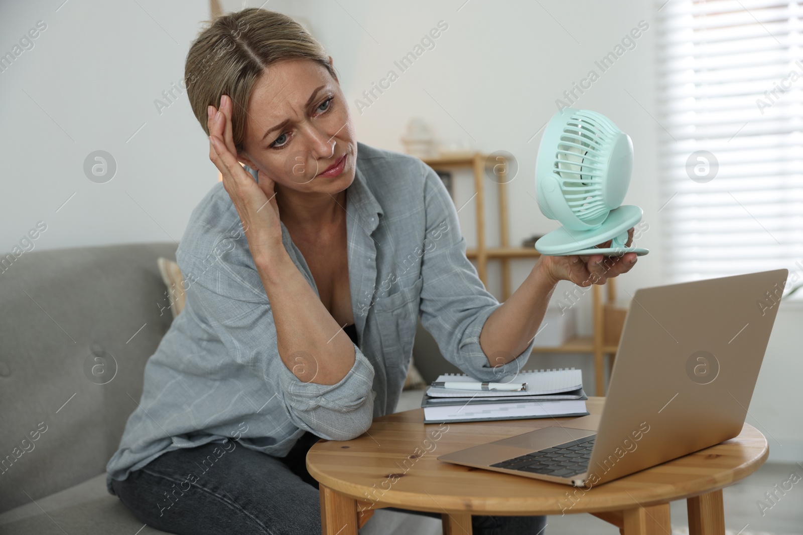 Photo of Menopause. Woman using electric fan to cool herself during hot flash at table indoors