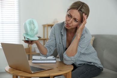 Menopause. Woman using electric fan to cool herself during hot flash at table indoors