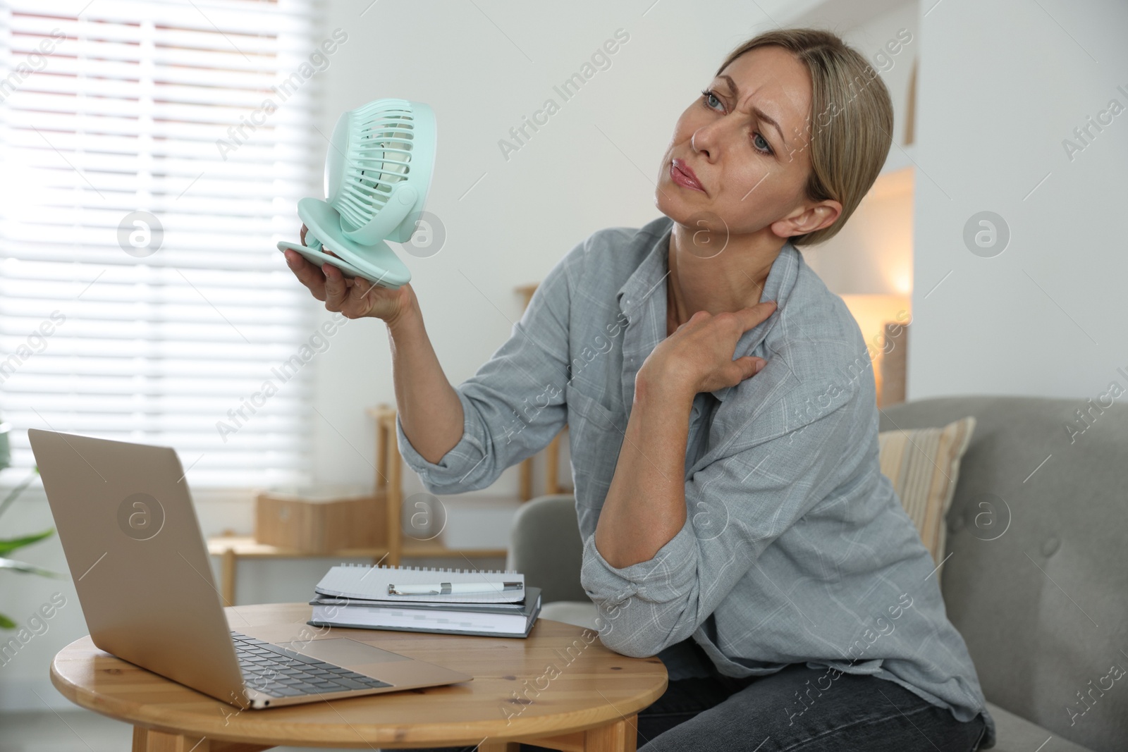 Photo of Menopause. Woman using electric fan to cool herself during hot flash at table indoors