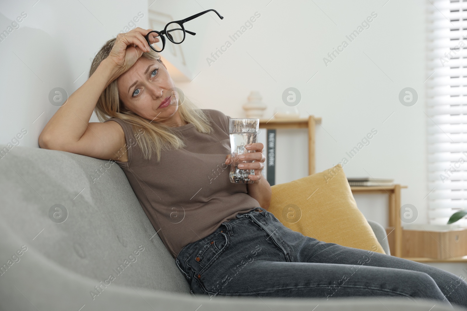 Photo of Menopause. Woman with glass of water suffering from headache on sofa at home