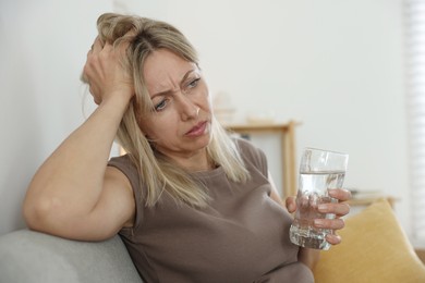 Photo of Menopause. Woman with glass of water suffering from headache on sofa at home