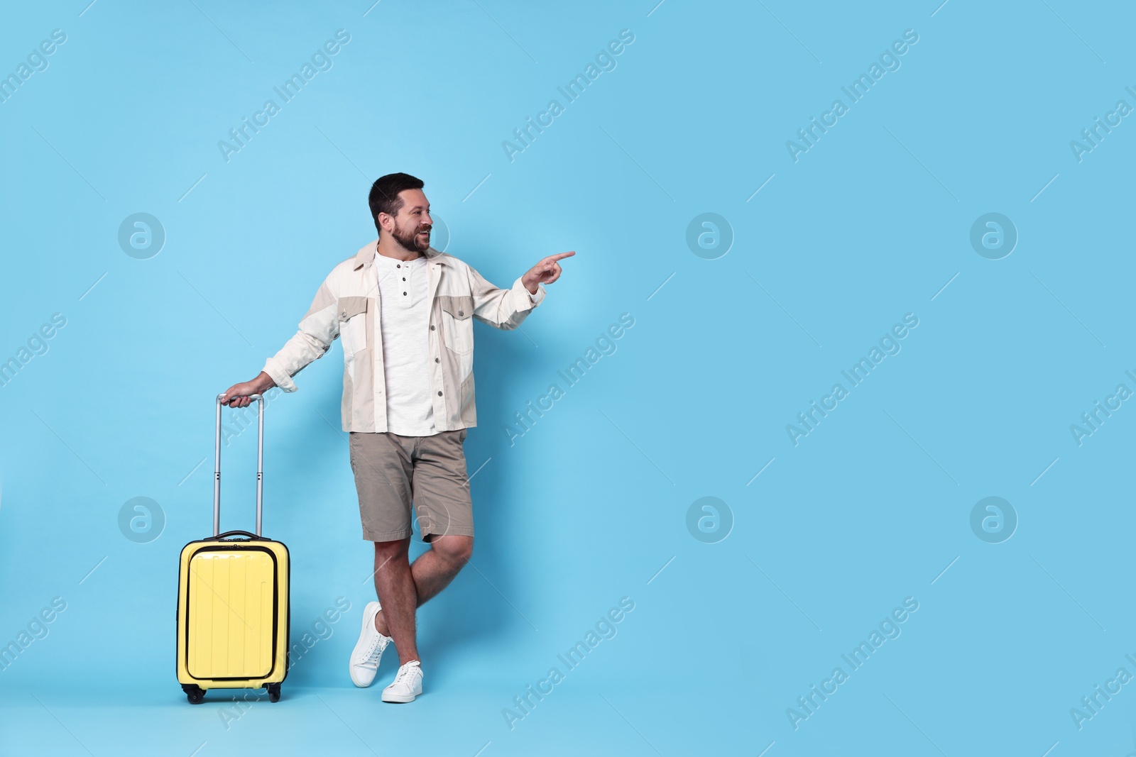 Photo of Happy man with suitcase pointing at something on light blue background, space for text