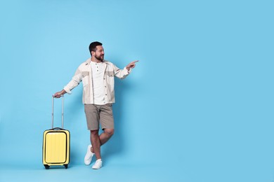 Photo of Happy man with suitcase pointing at something on light blue background, space for text