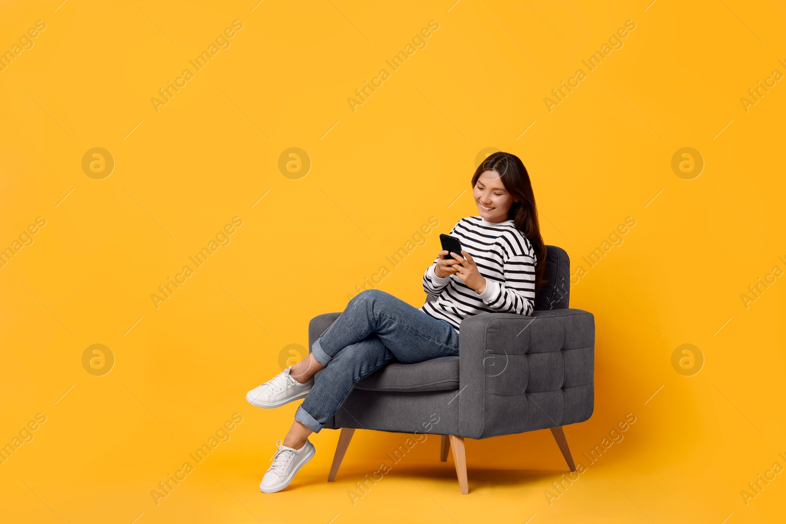 Photo of Smiling woman with smartphone sitting in armchair on yellow background. Space for text