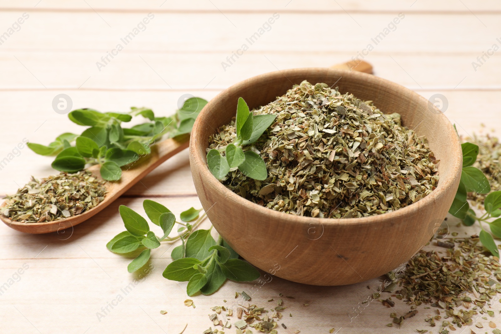 Photo of Dried oregano in bowl, spoon and green leaves on wooden table, closeup