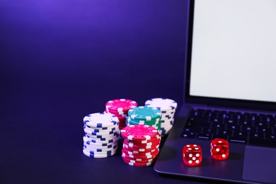 Photo of Online poker. Chips, dice and laptop on dark table, closeup