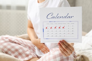 Photo of Woman holding calendar with marked menstrual cycle days on bed at home, closeup