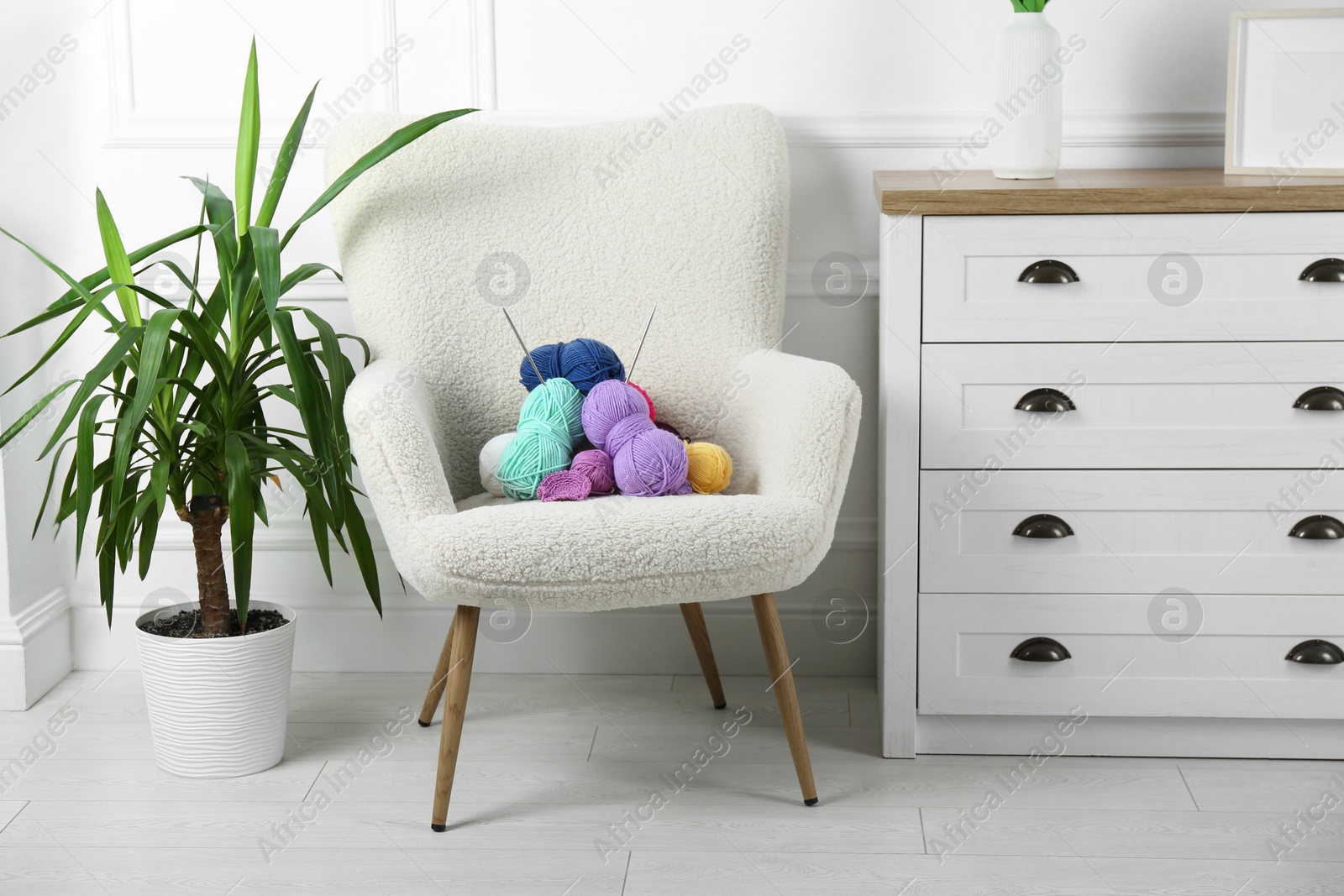 Photo of Armchair with pile of colorful yarns, crochet pattern and knitting needles between houseplant and chest of drawers indoors