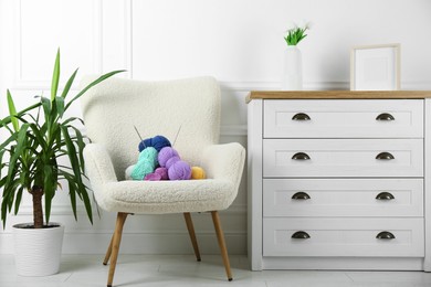 Photo of Armchair with pile of colorful yarns, crochet pattern and knitting needles between houseplant and chest of drawers indoors