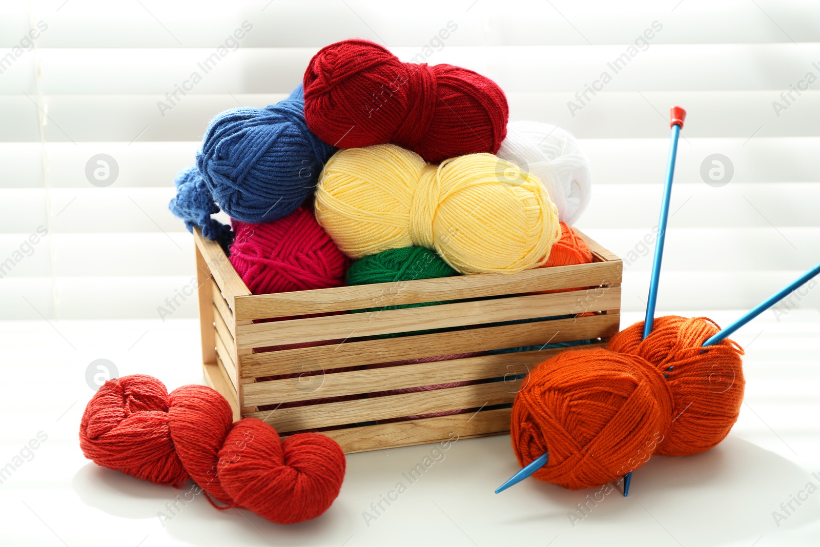 Photo of Skeins of soft yarn in crate and knitting needles on white table indoors