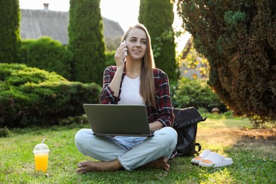 Photo of Woman with laptop talking on smartphone on green lawn in park