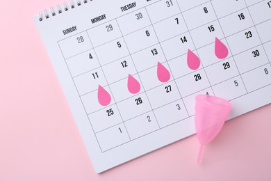 Photo of Calendar with marked dates and menstrual cup on pink background, top view