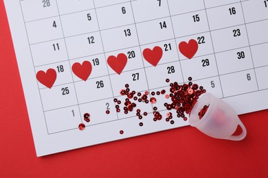 Photo of Calendar with marked dates and menstrual cup on red background, top view