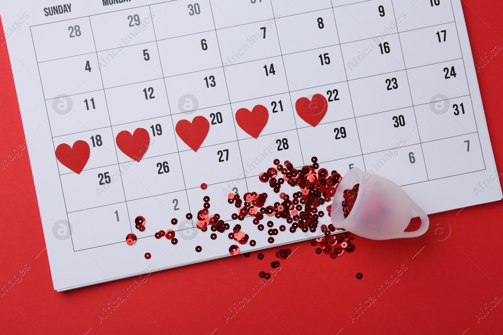 Photo of Calendar with marked dates and menstrual cup on red background, top view