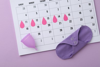 Photo of Calendar with marked dates, reusable pad and menstrual cup on violet background, top view