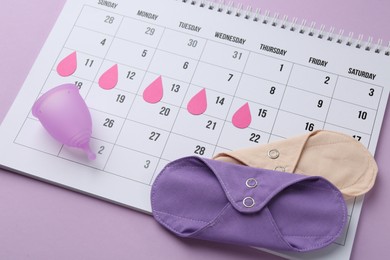 Photo of Calendar with marked dates, reusable pads and menstrual cup on violet background, closeup