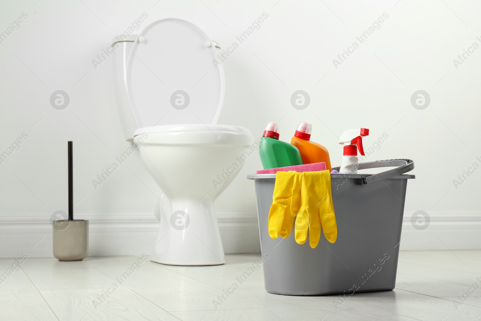 Photo of Bucket with different toilet cleaners and gloves on floor in bathroom