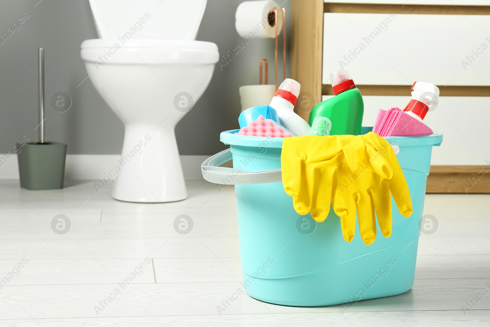 Photo of Bucket with different toilet cleaners, rag, sponge and gloves on floor in bathroom