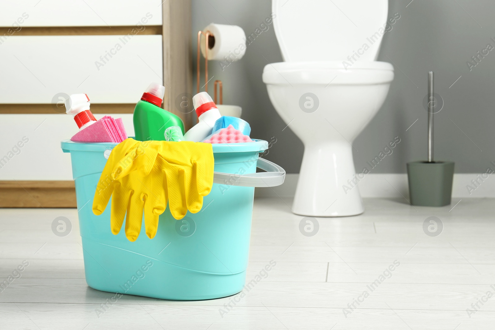 Photo of Bucket with different toilet cleaners, rag, sponge and gloves on floor in bathroom