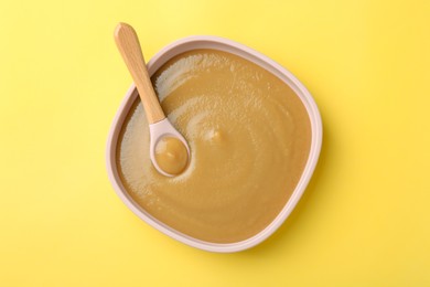 Photo of Delicious baby food with spoon in bowl on yellow table, top view