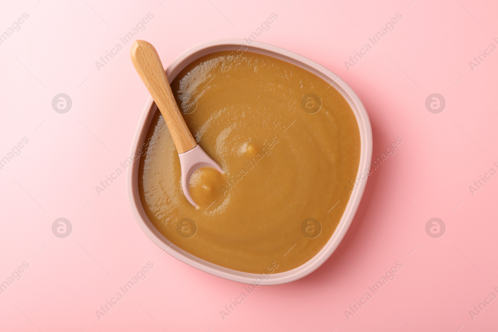 Photo of Delicious baby food with spoon in bowl on pink table, top view