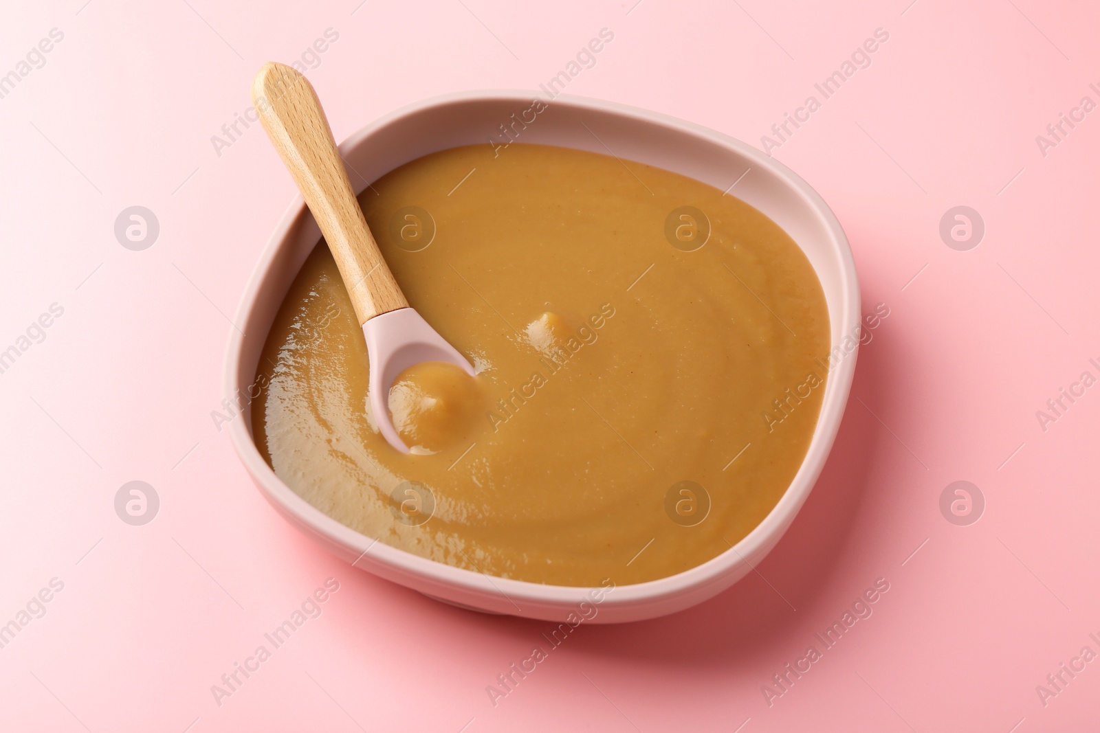 Photo of Delicious baby food with spoon in bowl on pink table