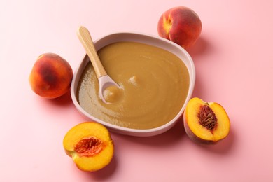 Photo of Delicious baby food with spoon in bowl and fresh peaches on pink table