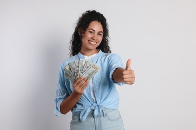 Happy woman with dollar banknotes showing thumbs up on light grey background