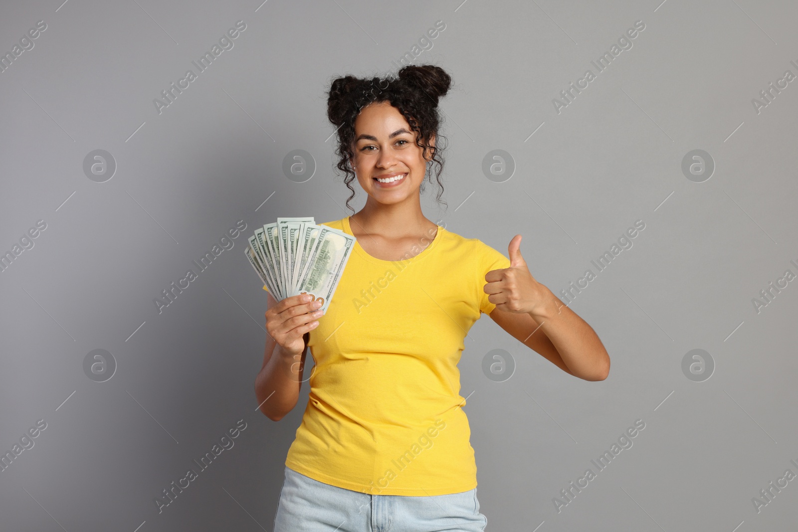 Photo of Happy woman with dollar banknotes showing thumbs up on grey background