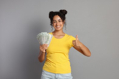 Happy woman with dollar banknotes showing thumbs up on grey background