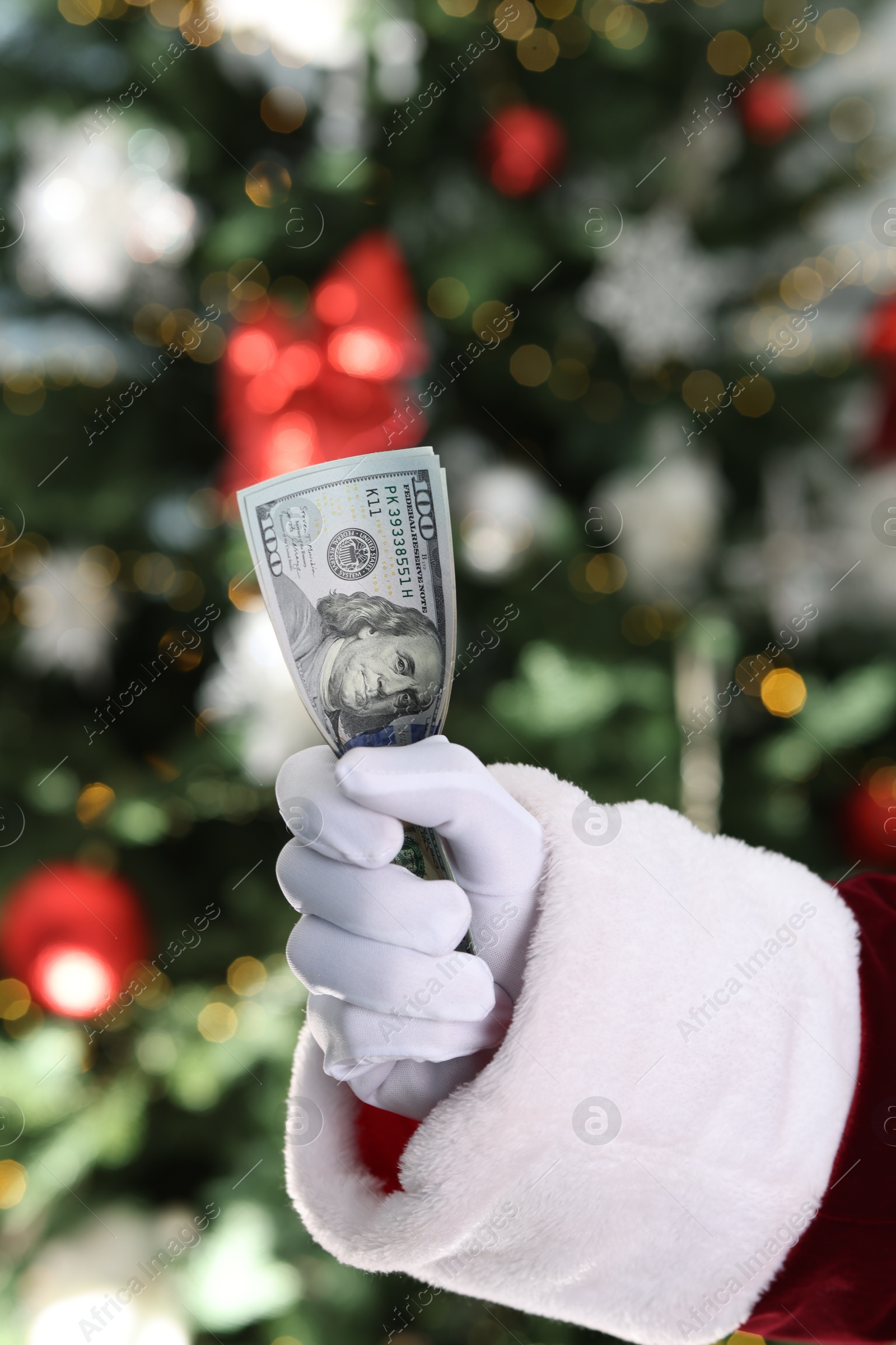 Photo of Santa Claus holding dollar banknotes against blurred Christmas lights, closeup