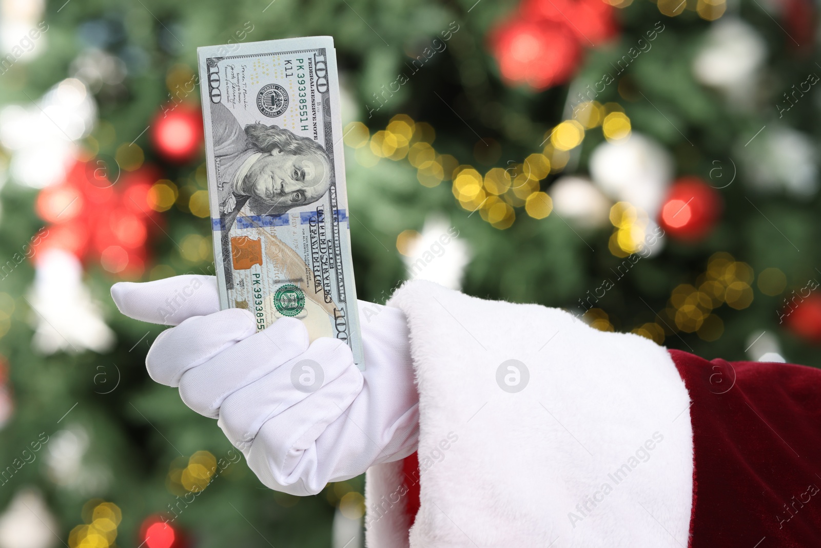 Photo of Santa Claus holding dollar banknotes against blurred Christmas lights, closeup