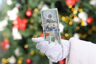 Photo of Santa Claus holding dollar banknotes against blurred Christmas lights, closeup