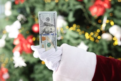 Photo of Santa Claus holding dollar banknotes against blurred Christmas lights, closeup