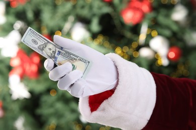 Photo of Santa Claus holding dollar banknotes against blurred Christmas lights, closeup