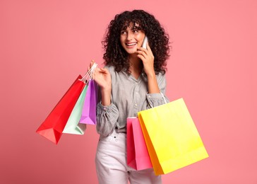 Smiling woman with colorful shopping bags talking by smartphone on pink background