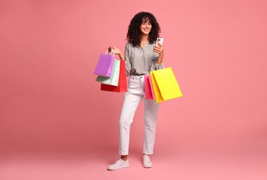 Smiling woman with colorful shopping bags and smartphone on pink background