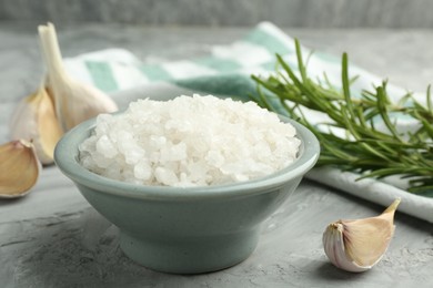 Photo of Sea salt in bowl, garlic and rosemary on gray textured table, closeup