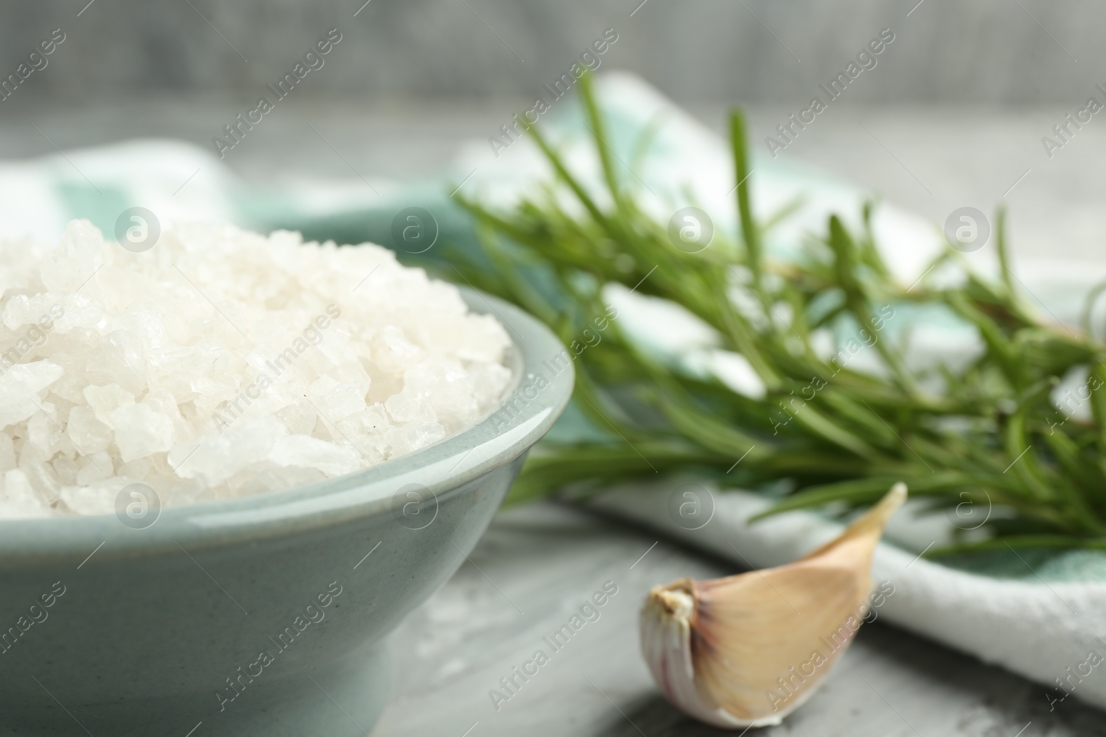 Photo of Sea salt in bowl, garlic and rosemary on gray table, closeup