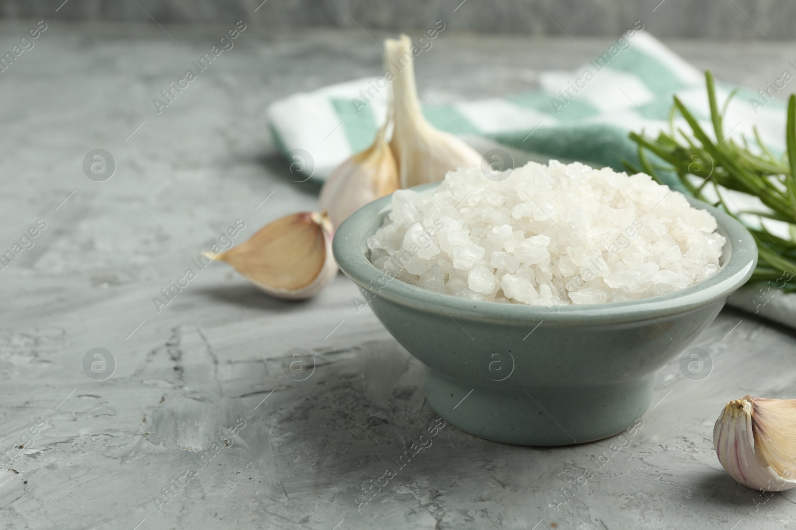 Photo of Sea salt in bowl, garlic and rosemary on gray textured table, closeup. Space for text