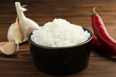 Sea salt in bowl, garlic and chili peppers on wooden table, closeup