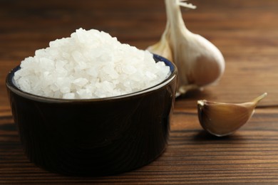 Photo of Sea salt in bowl and garlic on wooden table, closeup