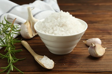 Sea salt in bowl, garlic and rosemary on wooden table, closeup