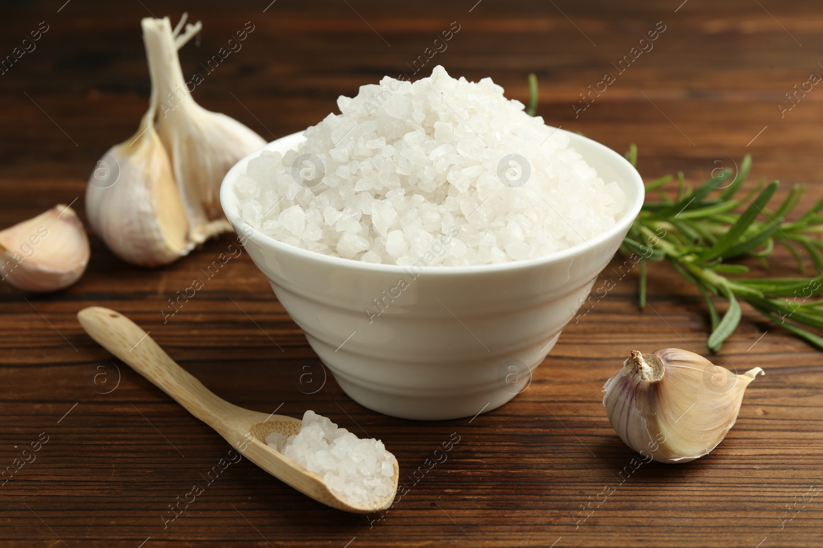 Photo of Sea salt in bowl, garlic and rosemary on wooden table, closeup