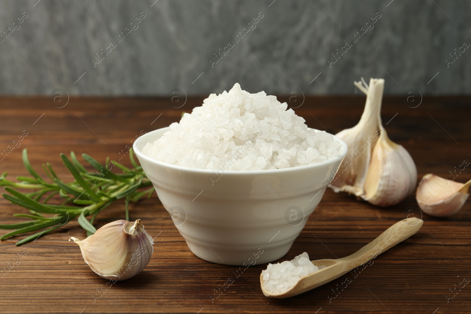 Photo of Sea salt in bowl, garlic and rosemary on wooden table, closeup