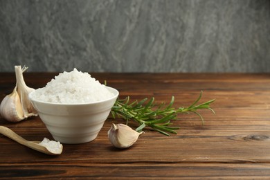 Photo of Sea salt in bowl, garlic and rosemary on wooden table. Space for text