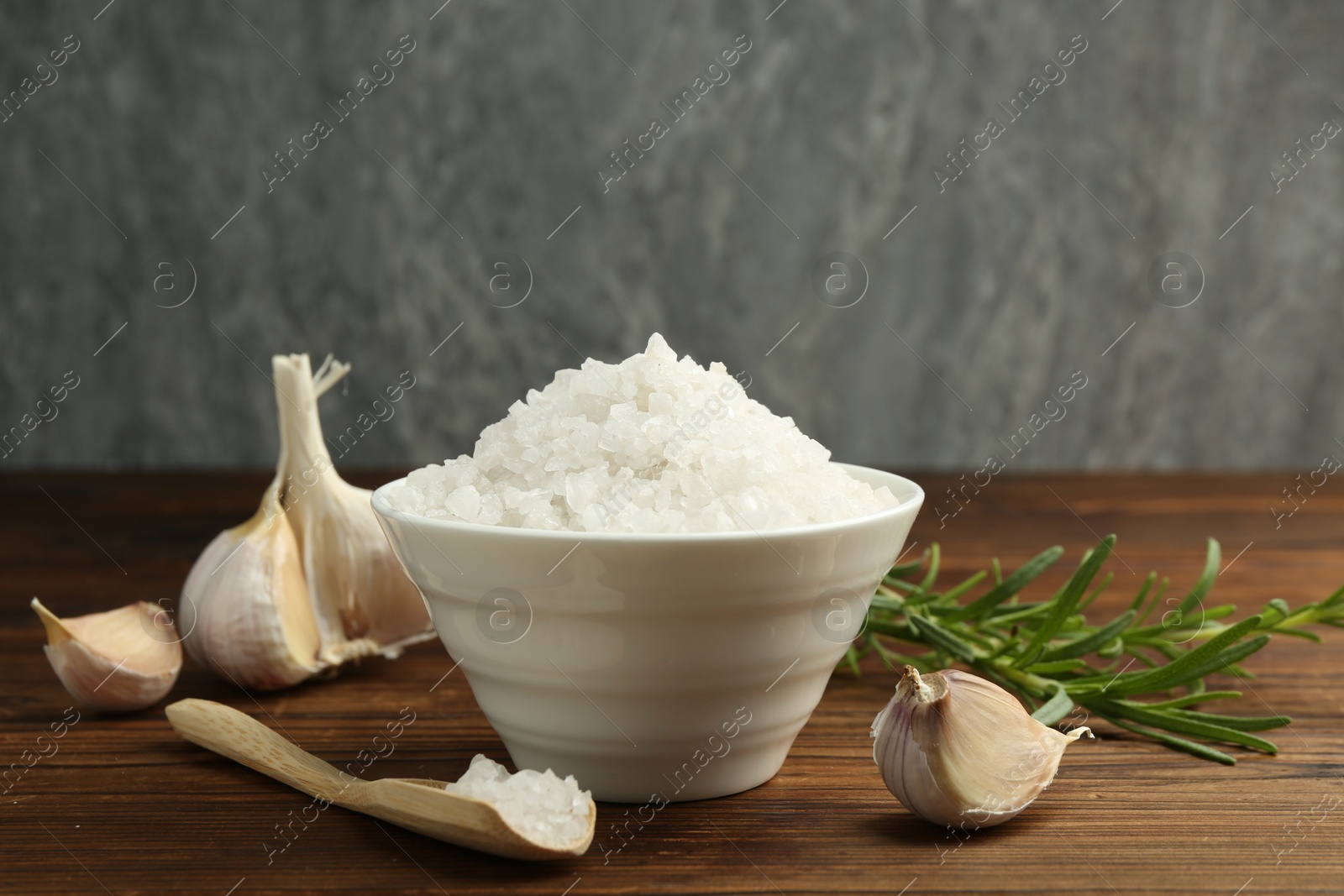 Photo of Sea salt in bowl, garlic and rosemary on wooden table