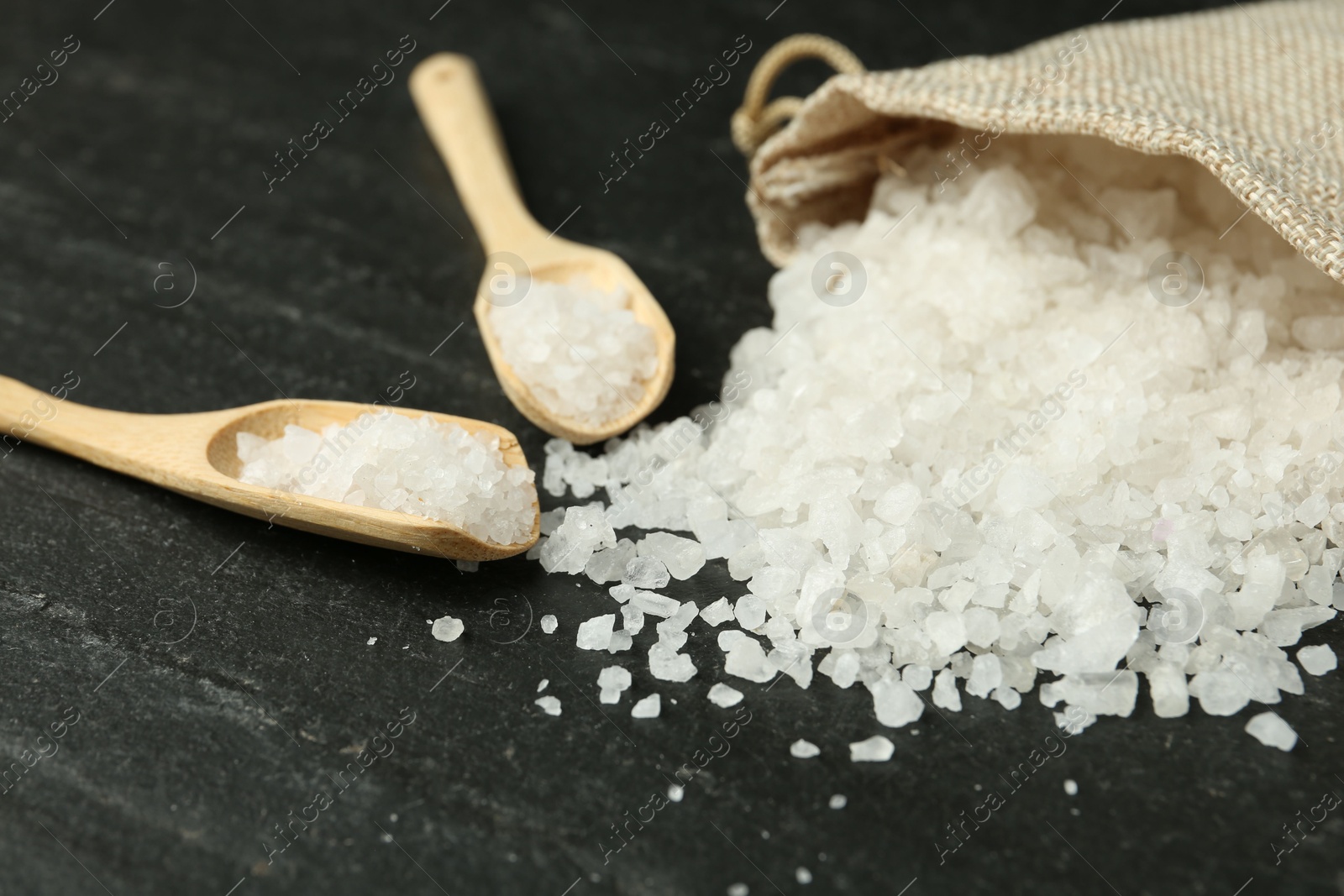 Photo of Sea salt, bag and spoons on dark gray textured table, closeup